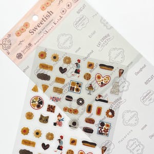 Puede incluir: Una hoja de pegatinas Sweetish con ilustraciones de postres, incluyendo galletas, macarons y éclairs. Diseñadas por Shinzi Katoh, las pegatinas incluyen las palabras "Cookies", "Doughnut" y "Biscuit".