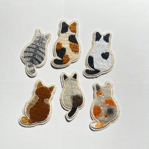 May include: Six embroidered iron-on patches shaped like cats viewed from behind. The collection includes a grey tabby, a calico, a white cat with black spots, a solid brown cat, a Siamese-style cat, and a multi-colored spotted cat.