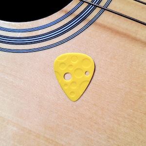 May include: A yellow guitar pick with a cheese-like design, featuring various sized circular cutouts. The pick is resting on the light wood surface of a guitar, with strings and the edge of the instrument visible.