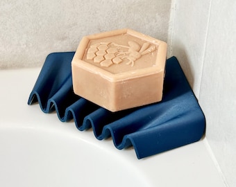 May include: A hexagonal beige soap bar with a bee and honeycomb design rests on a wavy, dark blue soap dish. The soap dish is placed in the corner of a white bathtub, providing a decorative and functional bathroom accessory.