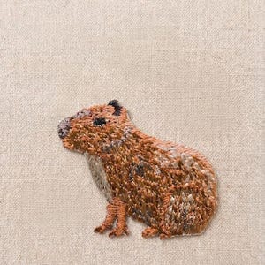 May include: Embroidered patch of a capybara in shades of brown, tan, and black. The capybara is depicted in a side profile, with detailed stitching creating a textured appearance. The patch is set against a neutral, textured background.