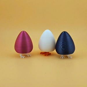 Puede incluir: Tres figuras en forma de huevo en magenta, blanco y azul marino. Cada huevo tiene pequeños pies blancos, con el huevo blanco con pies naranjas. Las figuras están sobre un fondo amarillo. Los huevos tienen una superficie texturizada.