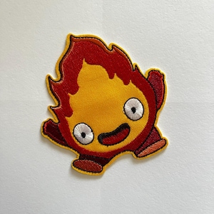 May include: An embroidered patch shaped like a stylized flame character with a bright yellow center, deep red outer edges, and a yellow border. The character features two large white circular eyes with black pupils and a small curved black mouth.