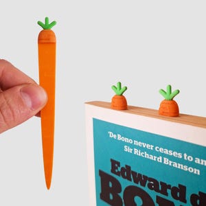 May include: Novelty carrot-shaped bookmarks in orange and green. One is held up, the others sit on a book's edge. The book has a teal cover with white text. A fun accessory for book lovers.