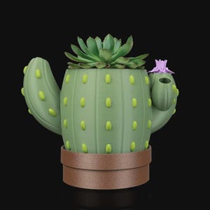 May include: A green cactus-shaped planter with a brown base, featuring yellow-green accents and a small purple flower. The planter holds a green succulent plant. The design is a unique decorative item for home or office.