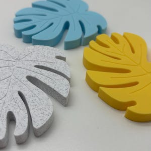 Puede incluir: Primer plano de tres posavasos con forma de hoja. Uno es azul claro, otro amarillo y el tercero blanco con motas grises. Los posavasos están hechos de un material sólido y tienen un tema tropical.