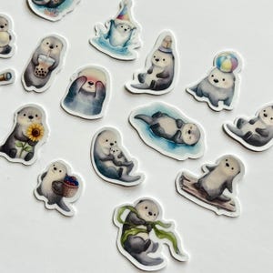 May include: A collection of die-cut stickers featuring various illustrations of sea otters. The otters are depicted in different poses, including holding a boba tea, a sunflower, a basket of berries, a beach ball, and wearing a party hat.