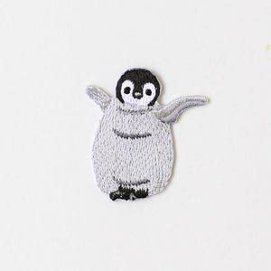 May include: Embroidered penguin patch with a black head, white face, and grey body. The penguin has its wings outstretched. The patch is made with detailed stitching, giving it a textured look. Suitable for attaching to clothing or accessories.