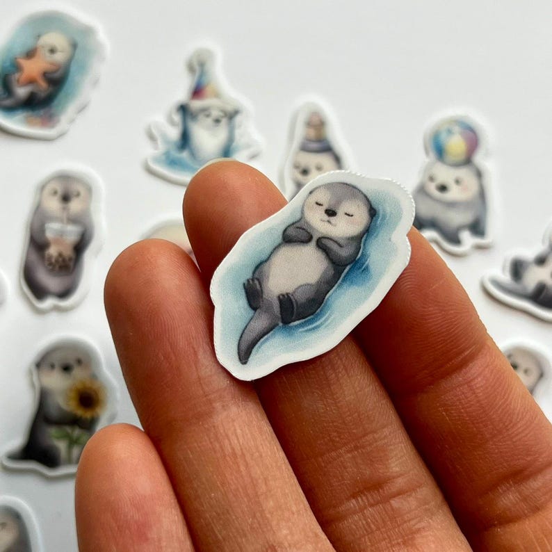 May include: A die-cut sticker featuring a gray and white sea otter floating on its back in blue water with its eyes closed. The otter has a rounded body, small paws tucked against its chest, and a dark tail. The sticker has a white border.