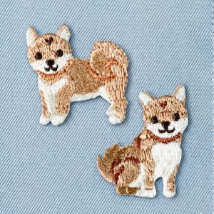 May include: Two embroidered Shiba Inu dog patches. The dogs are depicted in shades of brown, tan, and white, with black facial features and a red collar. The patches are on a light blue fabric background.