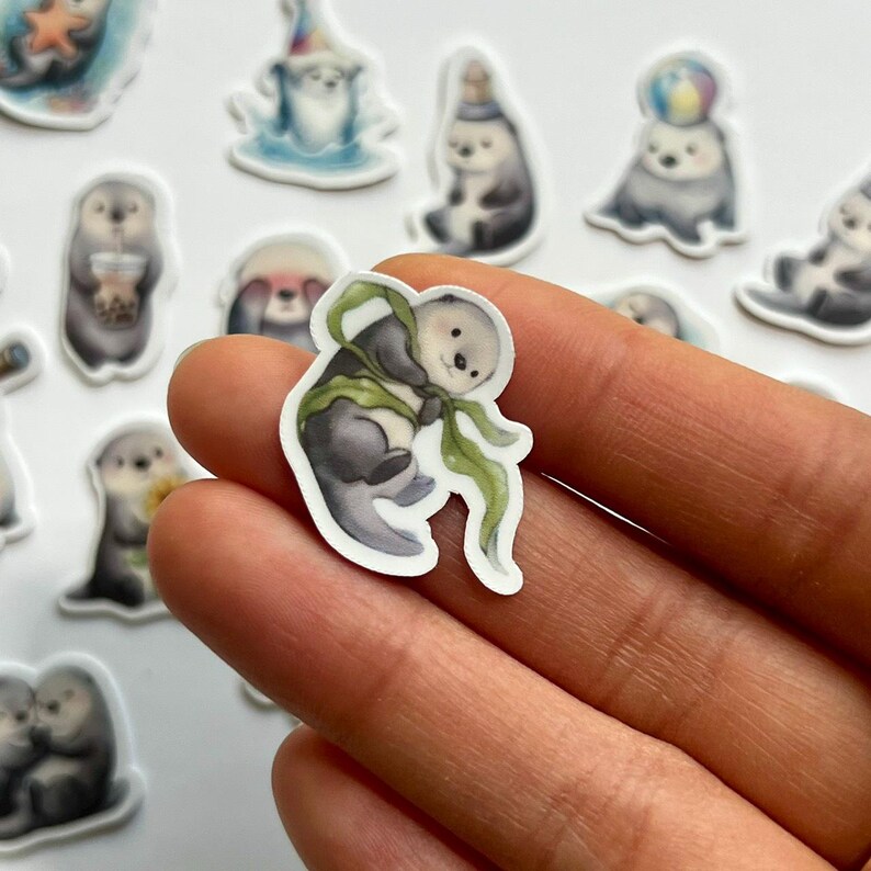 May include: A die-cut sticker featuring a watercolor-style illustration of a small sea otter wrapped in a long, flowing green kelp strand. The otter has a light-colored face, dark grey fur, and a curious expression, set against a white background.