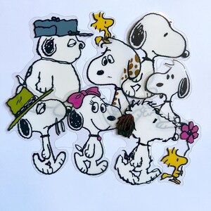 May include: A collection of clear stickers featuring cartoon dogs in various poses. The dogs are white with black accents, some wearing hats and bows. One sticker includes a yellow bird. The stickers are on a white background.