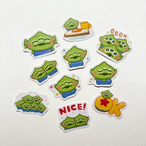 May include: A collection of ten die-cut stickers featuring green three-eyed alien characters in various poses and expressions. Some stickers include text such as OOOH, NICE!, and OK, while others show the aliens with food or in groups.