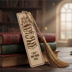Può includere: Segnalibro in legno con una pila di libri e un motivo floreale, con il testo "JUST ONE MORE PAGE". Ha un nappa oro e beige. Il segnalibro è appoggiato su una pila di libri con copertine rosse e verdi.