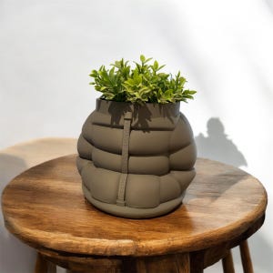 Pode incluir: Um vaso de plantas verde-oliva único em forma de casaco acolchoado, preenchido com folhagem verde vibrante. O vaso tem uma aparência acolchoada e um detalhe de alça. Está sobre uma mesa redonda de madeira, criando uma exibição moderna e elegante.