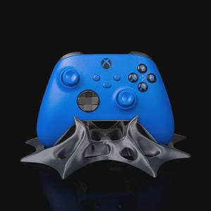 May include: A blue Xbox controller rests on a dark gray, stylized stand. The controller features the Xbox logo and various buttons. The stand has a unique, angular design, and the background is black.