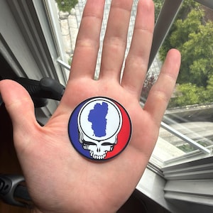 May include: A circular sticker featuring a stylized white skull graphic with a blue outline of a lake shape on the forehead, set against a background split into a blue section on the left and a red section on the right with a black border.