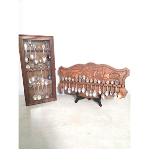 May include: A rectangular wooden display case with a glass front containing various metal souvenir spoons, positioned next to a carved wooden wall rack holding a row of decorative metal spoons with colorful emblems on their handles.