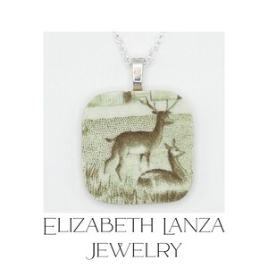 May include: A square-shaped pendant necklace featuring a green and white illustration of two deer in a field. The pendant is suspended from a silver-colored metal chain. Below the image, the text reads ELIZABETH LANZA JEWELRY.