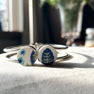 May include: Silver-tone metal cuff bracelet with two circular ceramic charms. Each charm features a white background with a blue floral or leaf design. The bracelet is resting on a soft, light-colored surface.