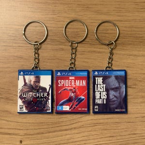 May include: Three keyrings with miniature video game box art. The keyrings have silver rings and chains. The games are "The Witcher 3: Wild Hunt", "Marvel Spider-Man", and "The Last of Us Part II". The game cases are blue with game art.
