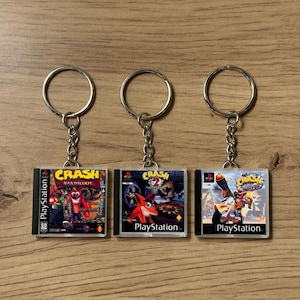 Könnte beinhalten: Drei Schlüsselanhänger mit Miniatur-PlayStation-Spielhüllen. Jeder Schlüsselanhänger zeigt ein anderes "Crash Bandicoot"-Spielcover, mit dem PlayStation-Logo. Die Schlüsselanhänger haben silberfarbene Schlüsselringe und Ketten.