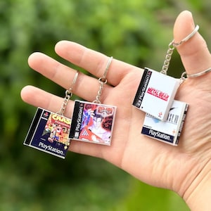 May include: Miniature PlayStation game case keyrings. The keyrings feature game covers from popular titles like Crash Bandicoot, Tekken 3, Metal Gear, and Final Fantasy VII. Each keyring has a silver-coloured chain and ring.