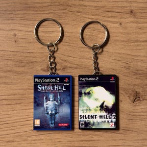 May include: Two keyrings featuring PlayStation 2 game covers. One shows "Silent Hill: Shattered Memories" with a child on a swing. The other displays "Silent Hill 2" with a ghostly image. Both have silver key rings.