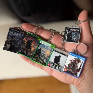 Peut inclure: Un ensemble de cinq porte-clés en forme de boîtiers de jeux vidéo miniatures avec les titres Fogbound, Worn, Horizon, Aftermath et Blacktide. Chaque boîtier rectangulaire en plastique présente une illustration détaillée, une chaîne et un anneau.