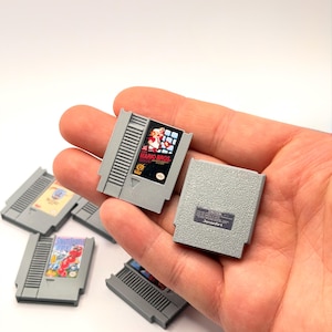 Puede incluir: Cartuchos de juegos de Nintendo grises, incluido uno con la etiqueta "Mario Bros." con un gráfico colorido. Los cartuchos están en una mano, con varios otros visibles en el fondo. Los cartuchos son rectangulares y tienen un diseño texturizado.