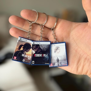 Può includere: Tre portachiavi a tema videogiochi. Ogni portachiavi presenta un design di custodia di gioco in miniatura con i titoli "The Last of Us Part I", "Resident Evil 4" e "Silent Hill 2". I portachiavi hanno catene e anelli color argento.