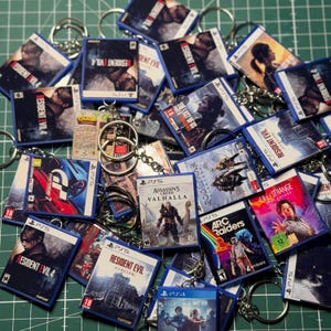 Könnte beinhalten: Eine Sammlung von Miniatur-Videospielhüllen-Schlüsselanhängern mit verschiedenen Titeln wie Resident Evil 4, Assassin's Creed Valhalla, God of War Ragnarok, Gran Turismo 7, Life is Strange True Colors und Arc Raiders, jeweils mit Metallringen.
