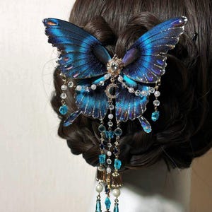 Puede incluir: Un accesorio para el cabello con una gran mariposa azul iridiscente con bordes oscuros. La mariposa est&aacute; adornada con un emblema met&aacute;lico central, peque&ntilde;as perlas blancas y m&uacute;ltiples cadenas colgantes con cuentas de cristal azul y peque&ntilde;as campanas doradas.