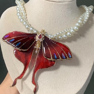 May include: A double-strand pearl necklace featuring a large butterfly pendant. The pendant has deep red wings with gold edging and iridescent blue details. A small, red gemstone is set in the centre of the pendant.