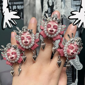 May include: A set of four unique rings featuring a pink face design with closed eyes, a skull crown, and dangling heart charms. The rings are adorned with pearls and small silver accents, creating a gothic and whimsical aesthetic.