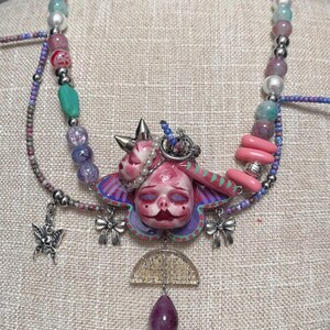 May include: A unique necklace featuring a central pendant with a pink, doll-like face and a butterfly-shaped design. The necklace incorporates various beads in shades of pink, purple, and blue, along with silver accents and a teardrop-shaped purple gem.