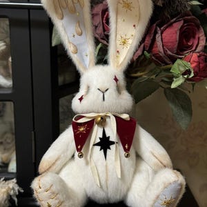 Puede incluir: Un conejo de peluche blanco con orejas largas y decoradas. Las orejas presentan detalles dorados y un dise&ntilde;o de goteo en una oreja y estrellas en la otra. Lleva un collar rojo y negro con una campana y una estrella. Las patas del conejo est&aacute;n bordadas con estrellas doradas.
