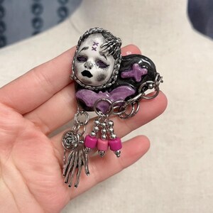 May include: A handcrafted brooch featuring a doll face with a black and silver color scheme. The brooch has a heart-shaped design with a purple cross and dangling pink beads. The brooch is held in a hand.
