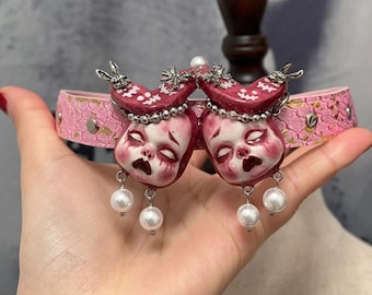 May include: A pink choker necklace featuring two doll-like faces with pearl accents. The faces have a distressed appearance with red and white coloring. The choker band is pink with a floral pattern and silver studs.