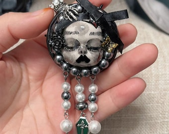 May include: A handcrafted brooch featuring a stylized face with closed eyes, a black mustache, and a crown. The brooch is adorned with black and gray beads, a black bow, and dangling pearl strands. A small green coffin charm hangs from the bottom.