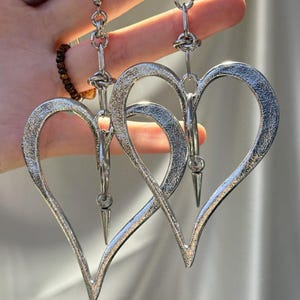以下が含まれることがあります： テクスチャード加工が施されたシルバーカラーのハート型イヤリング。各イヤリングは、ぶら下がっているスパイクペンダントが付いた大きなオープンハートのデザインが特徴です。イヤリングはチェーンとフックから吊り下げられています。