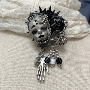 May include: A gothic-style brooch featuring a doll face, a heart with a cross, and dangling charms. The brooch is primarily black and silver, with accents of white and black beads. The doll face has stitched eyes and a sad expression.