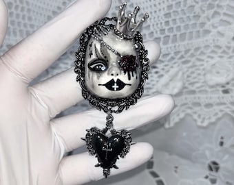 May include: A brooch featuring a pale, doll-like face with black markings, a silver crown, and a dark red eye detail, set within an ornate black frame. A black heart-shaped pendant with a small cross hangs from the bottom of the frame.