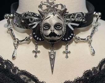 May include: Black leather choker necklace with a central pendant featuring a stylized cat face with a crown and butterfly wings. The choker has silver-tone chain accents, cross charms, and a dangling teardrop pendant. The cat face is painted in shades of gray and black.