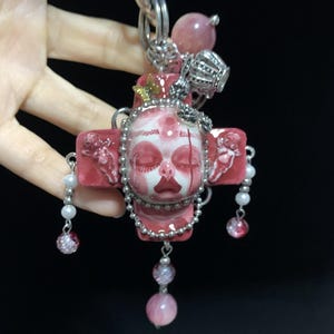 Puede incluir: Colgante rosa y blanco hecho a mano con un diseño de calavera estilizada. El colgante en forma de cruz presenta una calavera central, adornada con una corona plateada y detalles de cuentas. Cuentas rosas y transparentes cuelgan del colgante, creando un accesorio único.