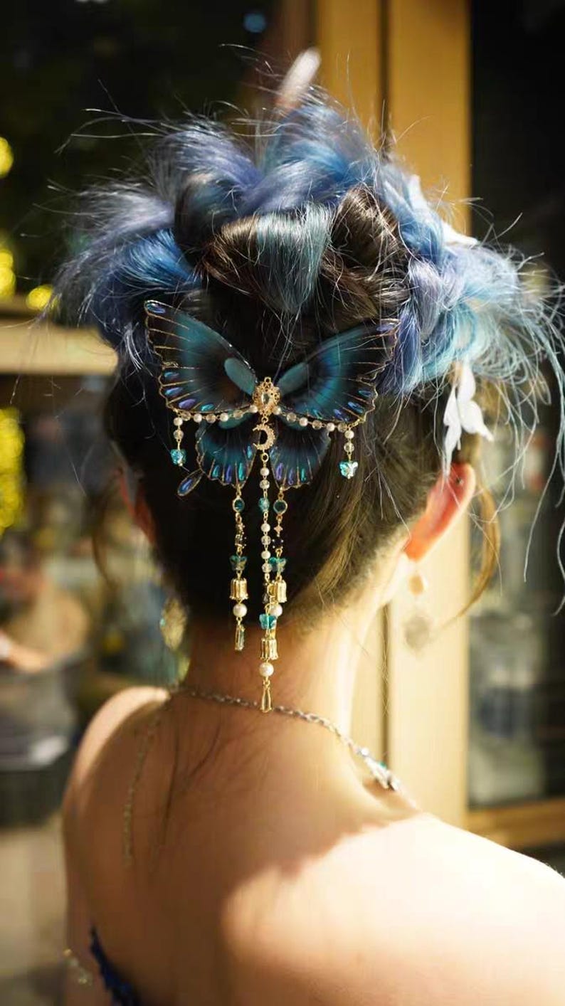Puede incluir: Un accesorio para el cabello con una mariposa grande de color azul oscuro con alas transl&uacute;cidas, detalles en metal dorado, un peque&ntilde;o dije de media luna y tiras colgantes de perlas blancas, cuentas transparentes y peque&ntilde;os dijes dorados.