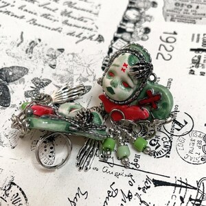 May include: A unique, handcrafted brooch featuring a doll-like figure with red eyes and a green face, adorned with a red cross and a silver skeletal hand. The brooch has a silver chain and green bead accents, with a ring attached. The background has vintage ephemera.