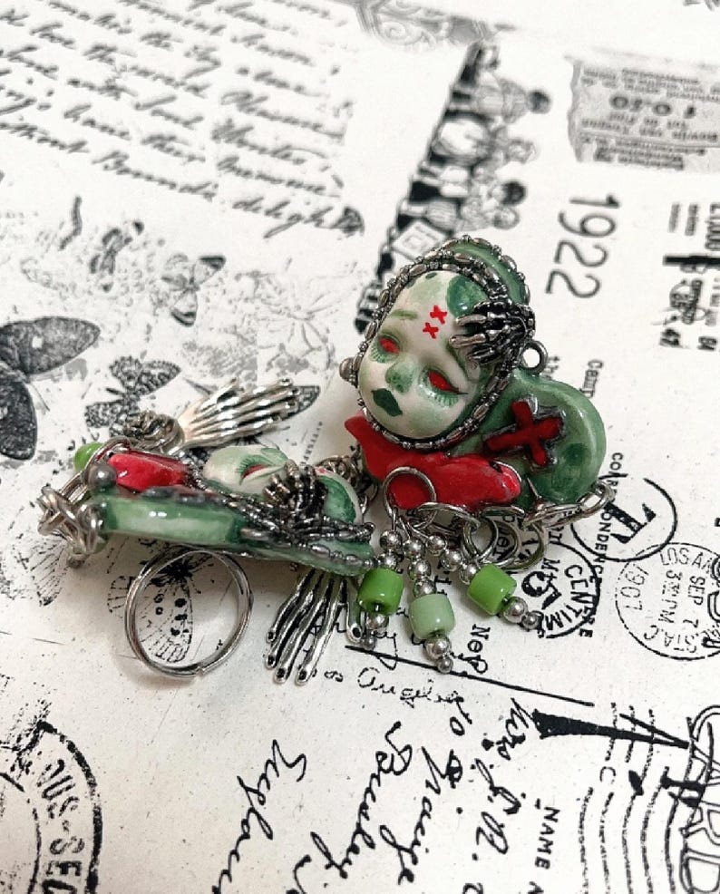 May include: A unique, handcrafted brooch featuring a doll-like figure with red eyes and a green face, adorned with a red cross and a silver skeletal hand. The brooch has a silver chain and green bead accents, with a ring attached. The background has vintage ephemera.
