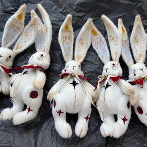 Puede incluir: Un grupo de mu&ntilde;ecos de peluche de conejos blancos con orejas largas. Las orejas est&aacute;n decoradas con dise&ntilde;os de velas. Cada conejo tiene una estrella roja en su cuerpo y un lazo rojo. Los mu&ntilde;ecos tienen una estrella negra en el pecho.