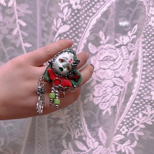 May include: A unique ring featuring a doll face with red eyes and a green face. The ring has a red bow, green beads, and a silver hand charm. The ring is held up against a white lace curtain.